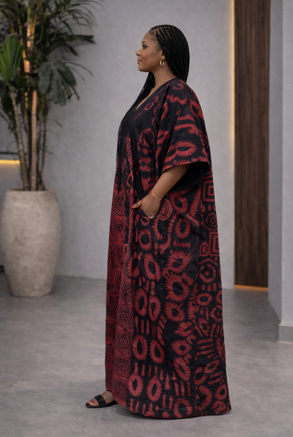 Woman wearing a red and black patterned dress standing indoors with a plant and door in the background.