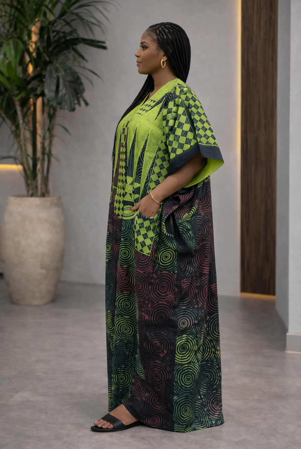 Woman wearing a green and black patterned outfit standing indoors with a plant in the background.