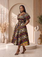 African print midi dress, African print dress UK, Ankara midi dress, puff sleeve African dress, African occasion dress, African wedding guest dress, fit and flare African dress, African wax print dress, contemporary African fashion, CUMO London dresses. A woman models a short puff-sleeve midi dress with a V-neck and a tied waist, featuring a vibrant multicolor geometric African wax print. She is standing in a styled studio setting with beige arches, wooden slats, and neutral decor.