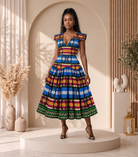 A woman models a vibrant, multicolored African print midi dress with a deep V-neckline and cap sleeves, standing on a round beige platform in a warm, sunlit studio setting.