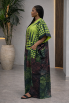Woman wearing a green and black patterned outfit standing indoors with a plant in the background.